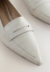 Valkoiset tekokrokotiili-nahasta valmistetut loafers-kengät, joissa on teräväkärkinen muotoilu, litteä pohja ja leveä nauha jalkapöydän yli, jossa on leikattu yksityiskohta.