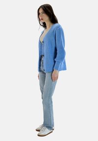 Lichtblauwe gebreide cardigan met lange mouwen, V-hals en knoopdetails, gedragen over een beige top, gecombineerd met lichtblauwe jeans en witte sneakers.