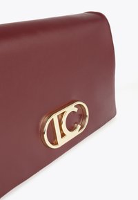 Burgundská kožená kabelka s výraznou zlatou sponou s logem, která předvádí hladké textury a elegantní design.