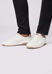 Sneakers bianchi in pelle con dettagli perforati, accenti beige sul tallone e suola in gomma texturizzata. Abbinati a pantaloni neri.