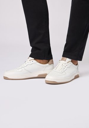 Weiße Ledersneaker mit durchbrochenen Details, beigen Akzenten an der Ferse und einer strukturierten Gummisohle. Kombiniert mit schwarzen Hosen.