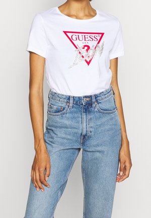 Kvinde iført en hvid Guess T-shirt med en rød trekantslogo og "Icon" skrevet i sølvfarve på lyseblå jeans med høj talje.