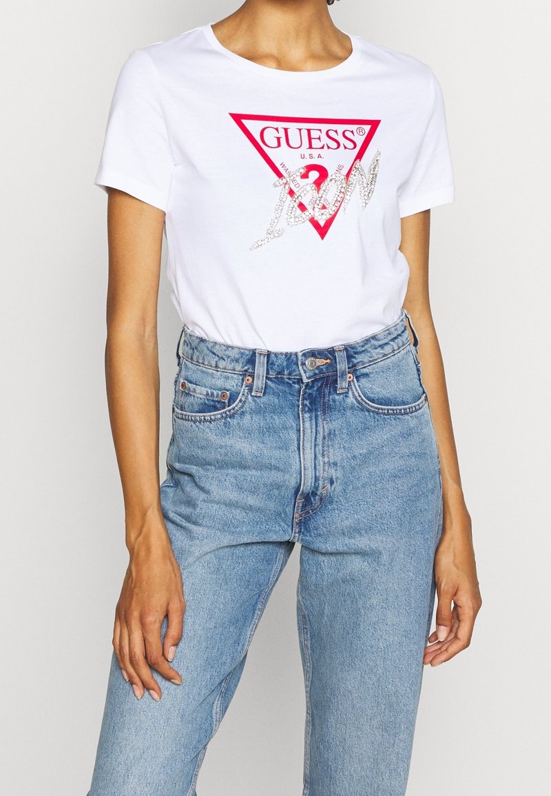 Femme portant un T-shirt blanc Guess avec un logo triangle rouge et "Icon" écrit en argent sur un jean bleu clair taille haute.