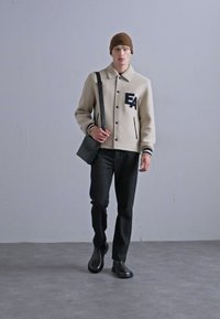 Jachetă din lână bej cu amestec de lână, cu dungi negre, având un logo "EA". Asortată cu pantaloni negri și o căciulă tricotată maro, ținând o geantă neagră.