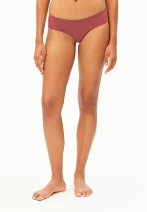Femme portant un bas de bikini mauve, debout pieds nus sur fond blanc, montrant les jambes et le bas du torse.