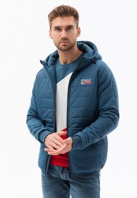 Giacca blu imbottita con zip e cappuccio, caratterizzata da pannelli in tessuto a contrasto, una piccola toppa con logo e polsini a costine. Indossata sopra una maglietta blu e rossa.