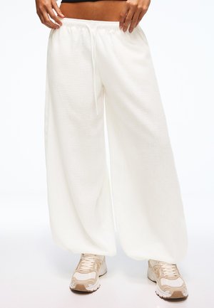 Osoba ubrana w luźne białe spodnie z troczkami, połączone z beżowo-białymi sportowymi butami, stojąca na jednolitym tle.