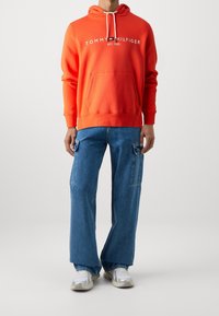 Sudadera naranja con bolsillo frontal y logo blanco, combinada con pantalones de mezclilla azul sueltos que tienen bolsillos laterales y zapatillas blancas.