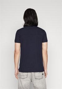 Man med axellångt mörkt hår som bär en marinblå kortärmad skjorta och ljusgrå jeans, vänd bort mot en enfärgad bakgrund.