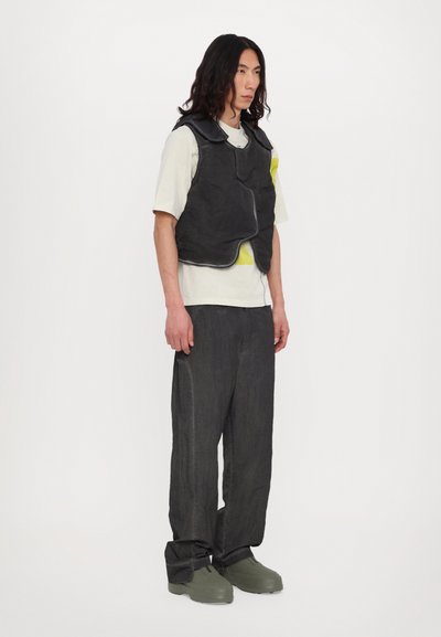 Gilet rembourré noir sur un t-shirt gris clair, associé à un pantalon gris foncé et des chaussures vertes imposantes. Textures lisses et design minimaliste.