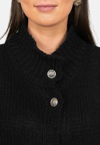 Zwart gebreid sweater met een hoge kraag, voorzien van twee zilveren knopen aan de voorkant. De textuur is dik en geribbeld.