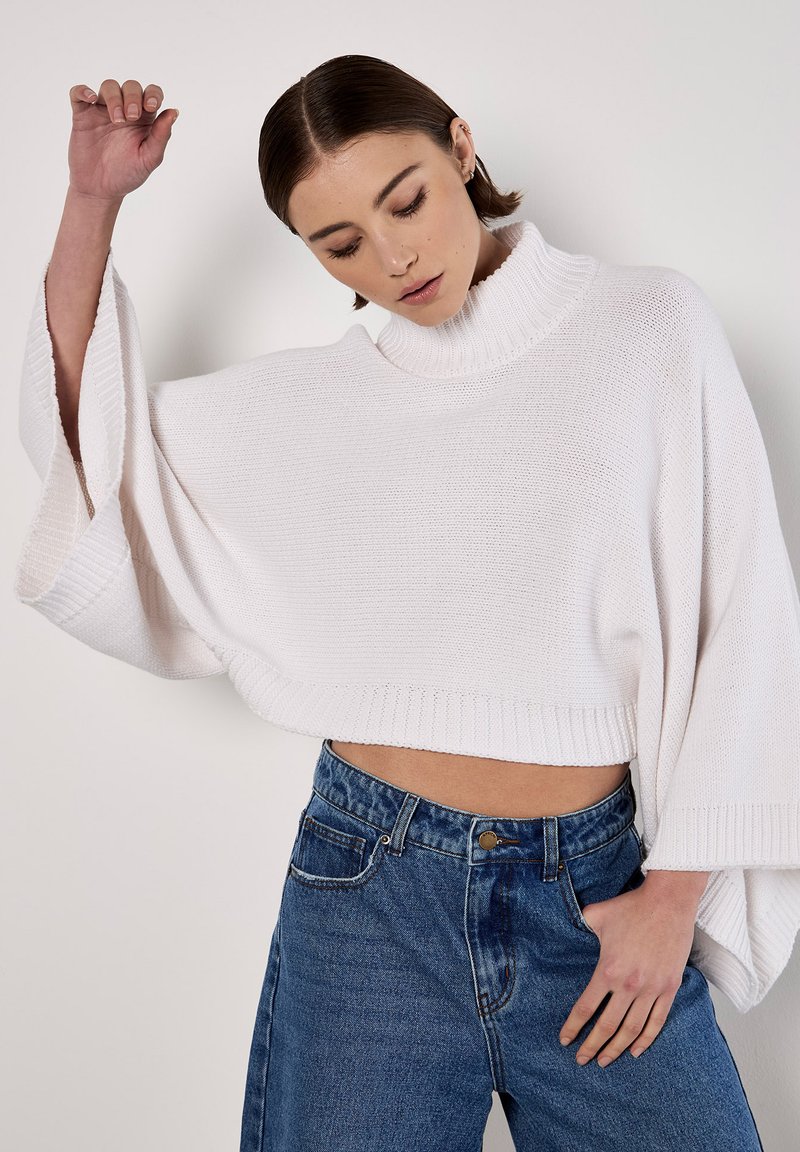 Weißer gestrickter, kürzerer Pullover mit hohem Kragen und weiten Ärmeln, kombiniert mit hochgeschnittenen blauen Jeans. Texturiertes Material, lässige Passform.