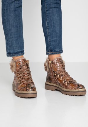 Bronsfärgade ankelstövlar med reptilmönster, pälsdetaljer, metallutrustning och snörning på en gummisula, kombinerade med blå jeans.