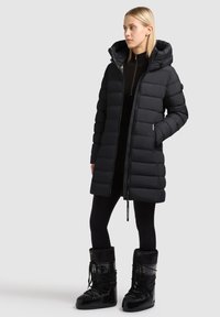 Black puffer coat with a hood, featuring a quilted design, front zipper, and side pockets. Worn over black leggings and boots.