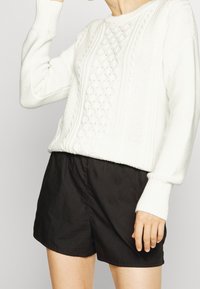 Biały dzianinowy sweter z teksturowanymi wzorami, o okrągłym dekolcie i długich rękawach, w połączeniu z czarnymi, luźnymi szortami.