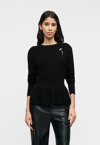 Μαύρο πλεκτό πουλόβερ με ριμπ ύφανση και πεπλούμ, μακριά μανίκια και στρογγυλή λαιμόκοψη· διαθέτει ένα μικρό μπλε λουλουδάτο καρφίτσα στην αριστερή πλευρά.