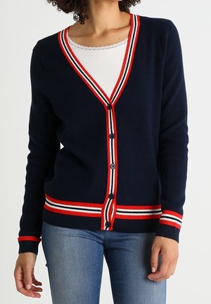 Vrouw draagt een navyblauw vest met rood, wit en zwart gestreepte bies over een wit topje met kanten rand en blauwe spijkerbroek, staat met ontspannen handen.