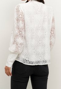 Vit spetsblus med blommönster, långa ärmar och rundad nederkant. Tyget är intrikat med utdragna detaljer och en lätt genomskinlighet.