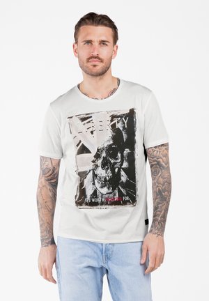 Mann mit Tattoos auf den Armen trägt ein weißes T-Shirt mit einem Totenkopf-Design und dem Text "Liberty" und "It's worth fighting for" sowie einer hellblauen Jeans.