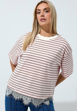 Giovane donna con lunghi capelli biondi che indossa una camicia a righe rosa e bianca con dettagli in pizzo e jeans blu, in piedi davanti a uno sfondo semplice.