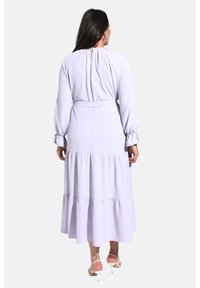 Vestido maxi de color púrpura claro con mangas largas, capas con volantes, cintura ceñida y detalle de escote en forma de lágrima en la parte posterior. Tejido suave con textura agradable.