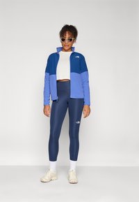 Kék és lila kapucnis dzseki magas gallérral, navy színű leggings-szel és krém színű tornacipővel párosítva. A dzsekin logó található a mellrészen.