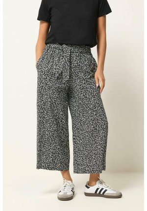 Woman wearing black patterned wide-leg cropped pants, black short-sleeve top, and white Adidas sneakers with black stripes.