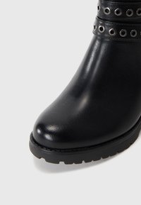 Black leather ankle boot with a rounded toe, textured finish, and decorative grommet accents on a wide strap. Thick rubber sole with tread.