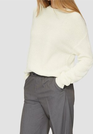 Personne portant un pull en maille blanc rentré dans un pantalon gris ample taille haute, les mains dans les poches, sur un fond gris clair.