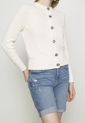 Cremfarbener Pullover mit strukturiertem Strickdesign und goldenen Knöpfen, kombiniert mit hochgekrempelten Jeansshorts mit angesagten Abnutzungsakzenten.