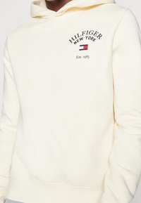 Felpa crema in tessuto morbido con tasca a marsupio. Presenta il testo "HILFIGER NEW YORK" e il logo in blu navy e rosso. Polsini e orlo a coste.