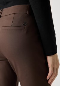 Detailní záběr na osobu, která má na sobě hnědé přiléhavé kalhoty s malým kovovým logem blízko zadní kapsy a černý dlouhý rukáv.