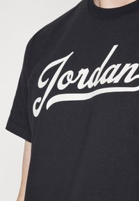 Černé tričko s krátkým rukávem s bílým kurzivním nápisem "Jordan" přes hruď, nošené osobou na jednolitém světlém pozadí.