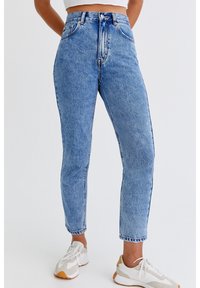 Jean en denim léger taille haute de couleur bleue, avec une coupe droite, présentant un léger effet usé, cinq poches et des coutures contrastées. Associé à des baskets blanches.