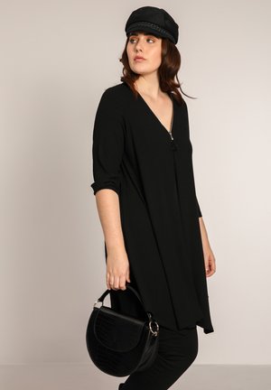 Schwarzes, geripptes Tunika mit V-Ausschnitt und Reißverschluss, ergänzt durch eine schwarze Handtasche im Krokodilmuster. Das Model trägt eine flache schwarze Mütze.