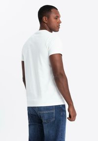 Camiseta blanca de manga corta con un cuello redondo, que presenta una textura suave. Llevada con jeans de mezclilla azul, mostrada desde un perfil lateral.