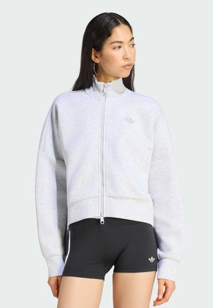 Žena u svijetlosivoj majici s patentnim zatvaračem i crnim sportskim kratkim hlačama s Adidas logotipima, gleda udesno na jednobojnoj pozadini.