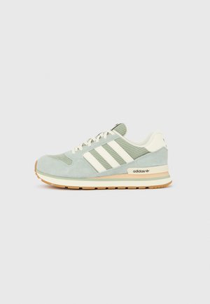 Baskets Adidas vert clair et beige avec des bandes blanches et des lacets, présentées sur un fond gris neutre, vue de profil.