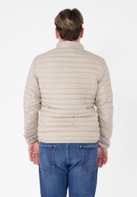 Giubbotto imbottito beige con trapuntatura, cuciture verticali, colletto alto e polsini elastici. Indossato sopra jeans blu, mostrato di spalle.
