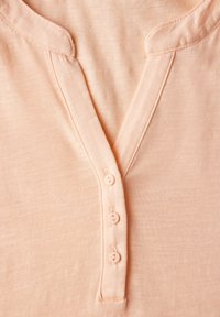 Top de color melocotón claro con un diseño de escote en V, tapeta de tres pequeños botones y tejido suave y texturizado. Costuras lisas a lo largo de los bordes.