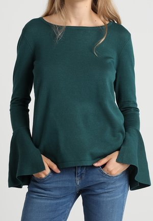 Femme portant un haut à manches longues vert foncé avec des poignets évasés et un jean bleu, les mains dans les poches, debout devant un fond uni.