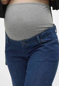 Sivi elastični maternični podkrajčnik nameščen pod modrimi jeans hlačami, ki imajo gumb in zadrgo ter naravni kroj.