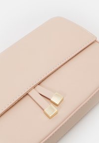 Portefeuille en cuir rose clair à texture lisse, avec une fermeture à rabat et deux accents rectangulaires dorés attachés à une sangle.