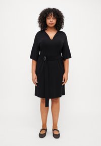 Černé šaty s opaskem, krátkými rukávy a výstřihem do v. Hladká látka, délka ke kolenům, opasek je zdobený kruhovou sponou. Kombinováno s černými balerínami.