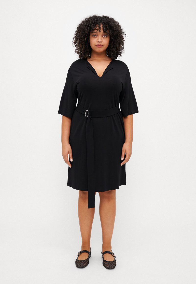 Černé šaty s opaskem, krátkými rukávy a výstřihem do v. Hladká látka, délka ke kolenům, opasek je zdobený kruhovou sponou. Kombinováno s černými balerínami.