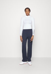 White long-sleeve athletic top with high collar. Navy blue wide-leg pants. Model wears white athletic shoes. Simple design, no visible patterns.