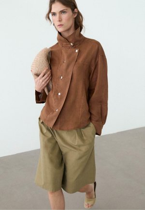 Donna con giacca marrone a collo alto con bottoni, pantaloncini larghi color oliva, sandali beige a ciabatta, che tiene in mano una clutch beige dalla trama evidente.