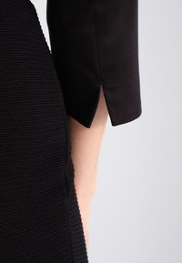 Černá texturovaná část oděvu se hladkým, přiléhavým rukávem s malým rozparkem na manžetě. Materiál je jemně žebrovaný a elegantní.