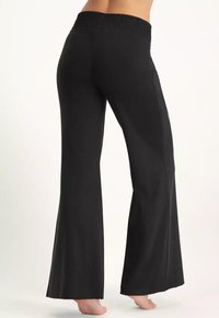 Person som bär svarta vida byxor med elastisk midja, står barfota vänd något åt sidan på en ljus bakgrund.