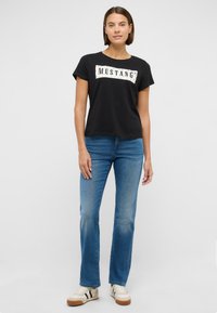 Melnas kokvilnas T-krekls ar “MUSTANG” grafiku, kombinēts ar zilām džinsa biksēm. Modelis valkā gaiļus sporta apavus ar tumšām svītrām un gumijas zoli.